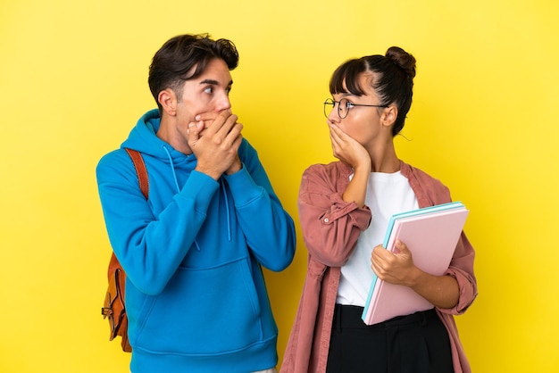 Giovane coppia di studenti isolata su sfondo giallo che copre la bocca con le mani per dire qualcosa di inappropriato