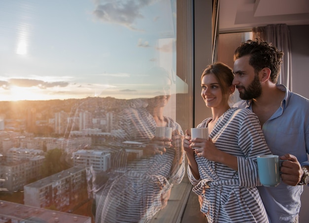 giovane coppia felice romantica che si gode il caffè serale e il bellissimo paesaggio al tramonto della città mentre si sta in piedi vicino alla finestra