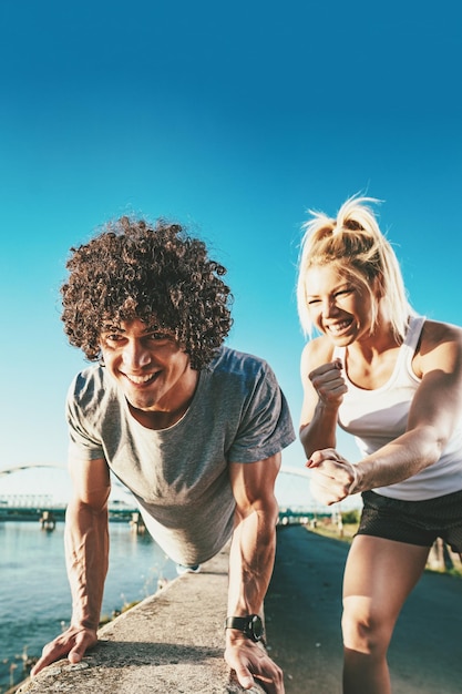 Giovane coppia fitness sta facendo allenamento sulla parete vicino al fiume in un tramonto. L'uomo è accovacciato e tiene in mano un kettlebell e la donna lo sostiene.