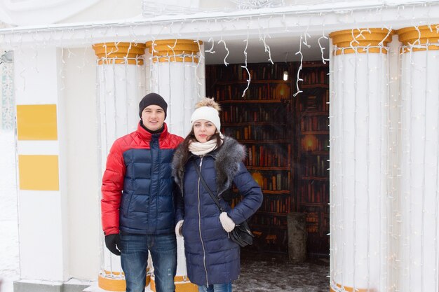 Giovane coppia in abiti invernali in posa all'aperto davanti a colonne bianche ornate che fiancheggiano un ingresso decorato con una ghirlanda di piccole feste o luci di Natale