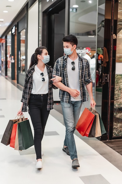 Giovane coppia in maschera di protezione che tiene più shopping bag di carta che cammina nel corridoio di un grande centro commerciale