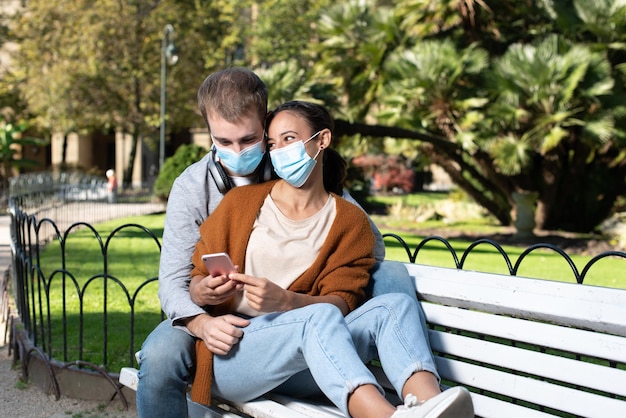 Giovane coppia in posizione romantica seduta su una panchina Amanti che controllano un telefono indossando maschere facciali durante la pandemia di Covid19 in un parco