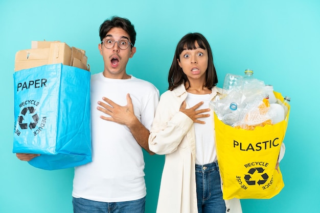 Giovane coppia in possesso di un sacchetto pieno di plastica e carta da riciclare isolato su sfondo blu sorpreso e scioccato mentre guarda a destra
