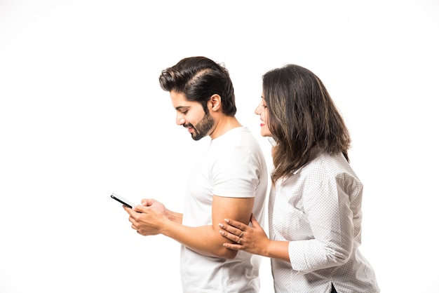 Giovane coppia indiana che utilizza smartphone o telefono cellulare, in piedi isolato su sfondo bianco o su sfondo di mattoni rossi
