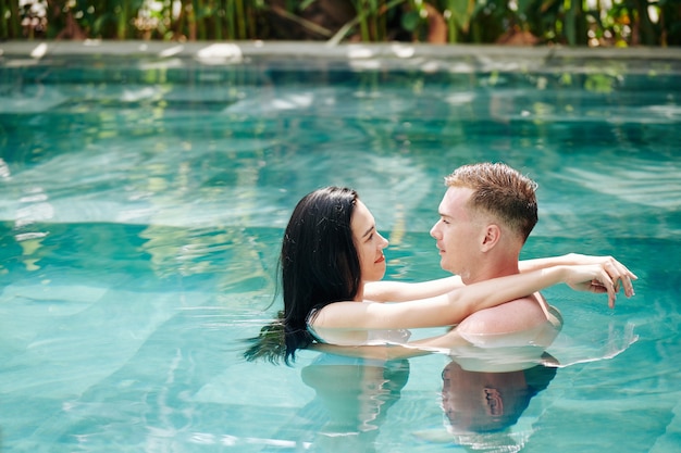 Giovane coppia innamorata in piedi in piscina, abbracciarsi e guardarsi l'un l'altro