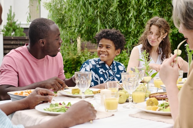 Giovane coppia interculturale che guarda il loro simpatico figlio che parla con suo padre mentre è seduto al tavolo servito tra i suoi genitori e si gode la cena