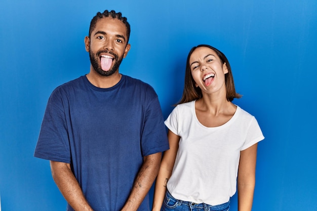 Giovane coppia ispanica in piedi insieme che attacca la lingua fuori felice con il concetto di emozione di espressione divertente