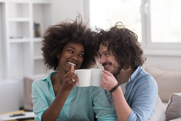 giovane coppia multietnica seduta sul divano di casa a bere caffè, parlare, sorridere.