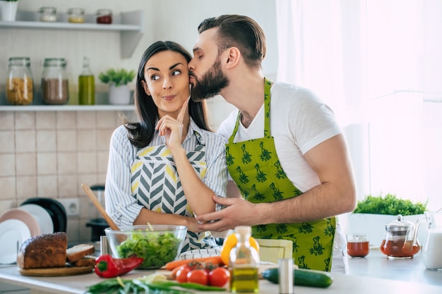 Giovane coppia sorridente eccitata innamorata che prepara un'insalata vegana super sana con molte verdure in cucina e il marito che bacia la moglie