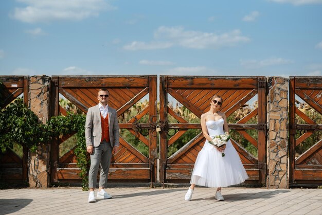 Giovane coppia sposa in un abito corto bianco e sposo in un abito grigio vicino al cancello di legno