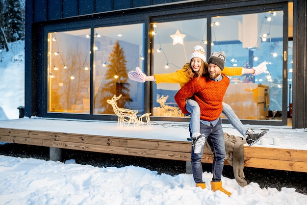 Giovane coppia vestita con maglioni e cappelli luminosi che si divertono insieme vicino alla casa decorata durante le vacanze invernali