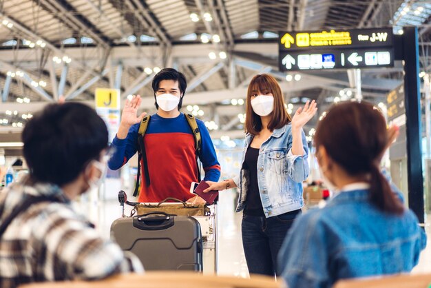 Giovane coppia viaggiatrice in quarantena per coronavirus che indossa una maschera chirurgica protezione per il viso salutando e salutando con un amico prima di un lungo volo di vacanza all'aeroporto internazionale del terminal