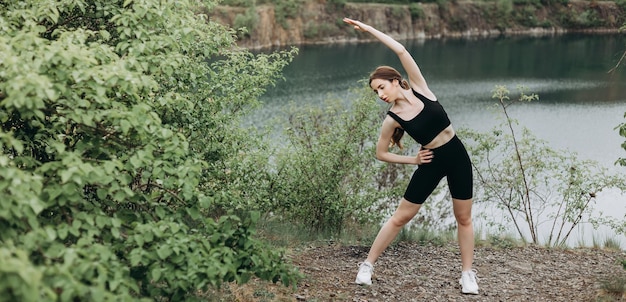 Giovane corridore femminile che allunga le braccia e le gambe prima di correre al sentiero forestale mattutino vicino al lago