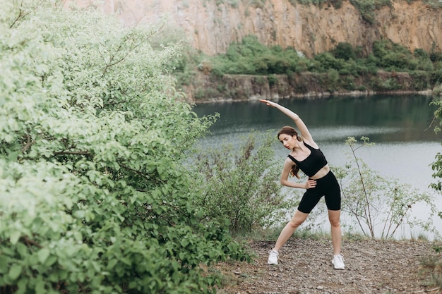 Giovane corridore femminile che allunga le braccia e le gambe prima di correre al sentiero forestale mattutino vicino al lago