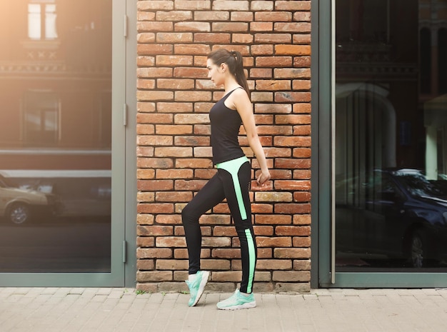 Giovane corridore femminile che si riscalda prima di correre, allungando le mani, sfondo del muro di mattoni, spazio per la copia