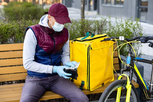Giovane corriere in maschera medica che consegna cibo con zaino termico giallo, in bicicletta in città. Concetto di servizio di consegna cibo.