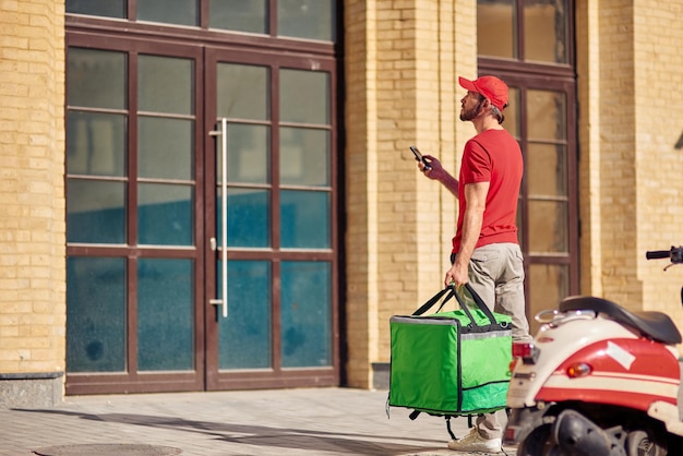 Giovane corriere maschio in uniforme rossa con borsa termica che usa il suo smartphone mentre si trova vicino alla casa dei clienti sulla strada. Servizi di consegna cibo