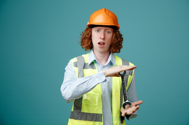 Giovane costruttore in uniforme da costruzione e casco di sicurezza che mostra la chiave inglese che sembra stupito e sorpreso in piedi su sfondo blu