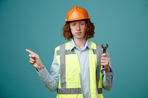 Giovane costruttore in uniforme da costruzione e casco di sicurezza che tiene la chiave che punta con il dito indice di lato guardando fiducioso in piedi su sfondo blu