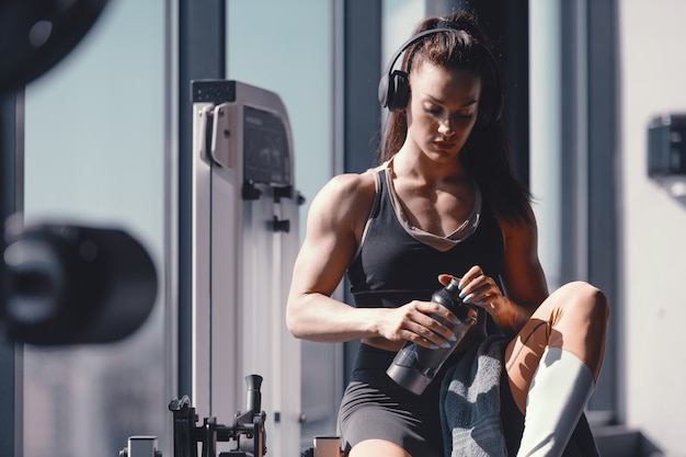 Giovane culturista femminile muscolare forte caucasico che si siede in ginnastica con le cuffie sulle orecchie e sull'acqua in mani. Soffri il dolore della disciplina di soffrire il dolore del rimpianto.