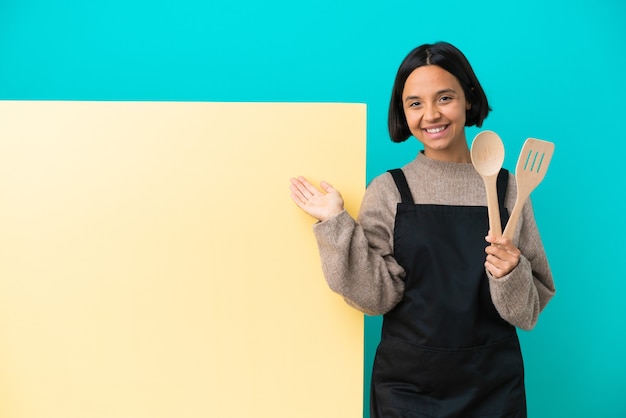 Giovane cuoca di razza mista con un grande cartello isolato su sfondo blu che allunga le mani di lato per invitare a venire