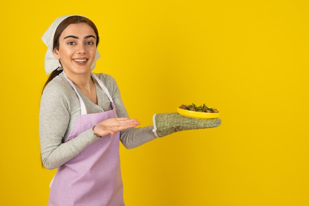 Giovane cuoca in grembiule che tiene piatto di funghi fritti sul muro giallo.