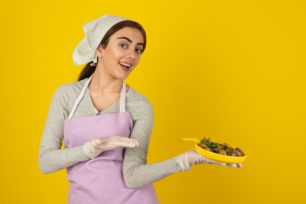 Giovane cuoca in grembiule che tiene piatto di funghi fritti sul muro giallo.