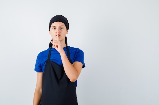 Giovane cuoco adolescente in t-shirt, grembiule che mostra gesto di silenzio e aspetto misterioso, vista frontale.