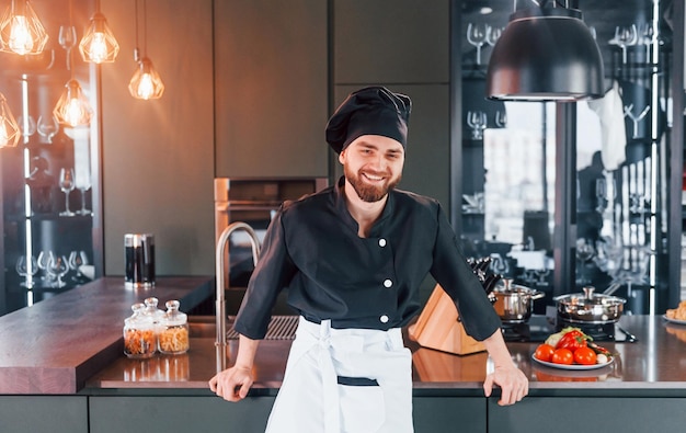 Giovane cuoco professionista in uniforme in piedi vicino al tavolo e in posa per una macchina fotografica in cucina