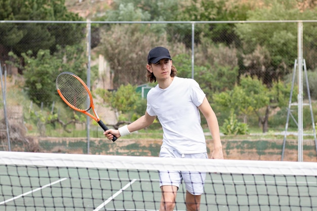 Giovane dai capelli lunghi con un berretto che gioca una partita di tennis su un campo con la rete nel mezzo Cercando di colpire la palla