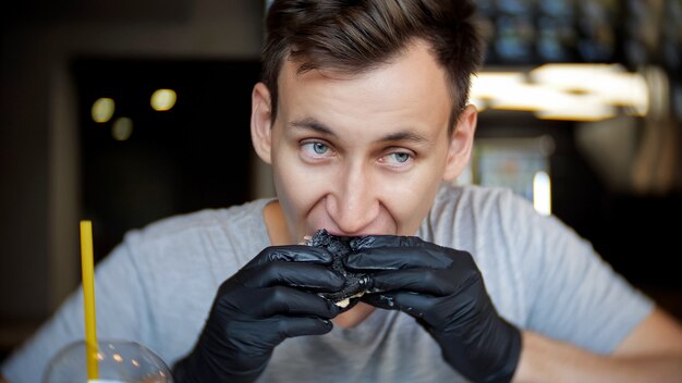 Giovane dell'uomo in guanti neri che mangia un hamburger in un caffè.