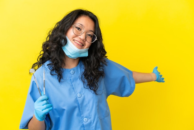 Giovane dentista asiatico che tiene gli strumenti su sfondo isolato estendendo le mani di lato per invitare a venire