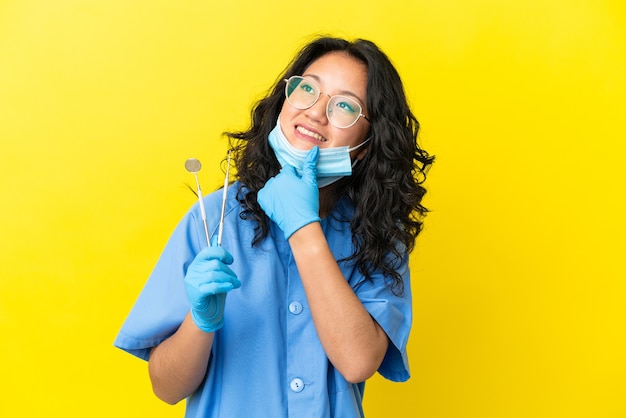 Giovane dentista asiatico che tiene gli strumenti su sfondo isolato guardando di lato e sorridente