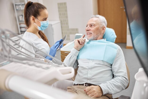 Giovane dentista professionista che tiene in mano un tablet e guarda un uomo anziano su una poltrona del dentista durante una consultazione