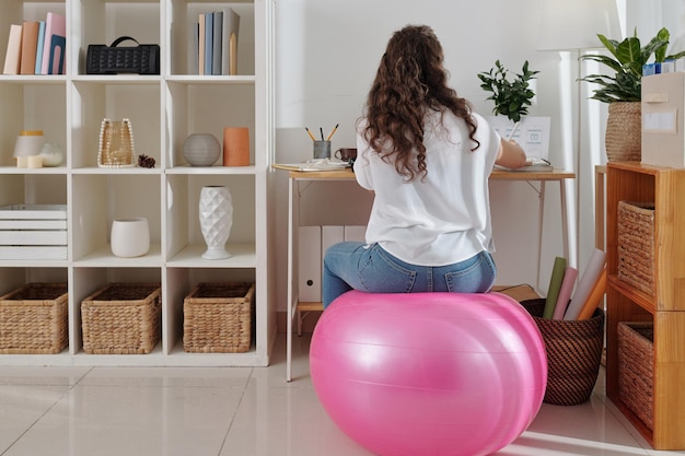 Giovane designer dell'interfaccia utente femminile seduta alla palla fitness quando si lavora su un progetto nel suo ufficio a casa