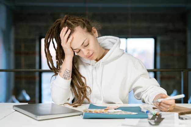 Giovane designer femminile stanco che tocca la testa mentre si china sulla scrivania e sceglie il colore per la nuova collezione