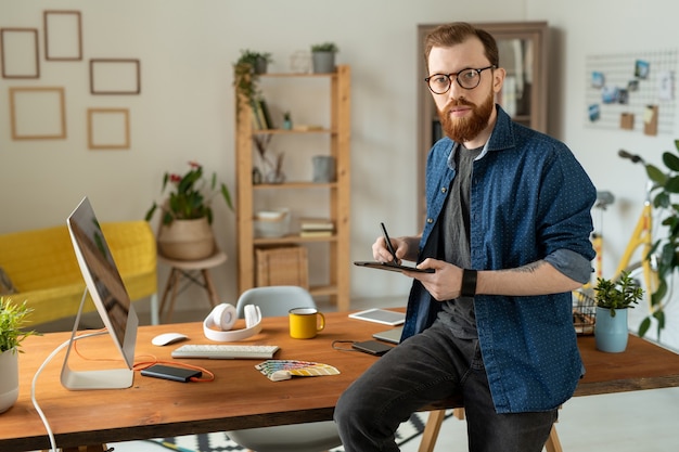 Giovane designer freelance barbuto che tiene in mano uno stilo e un tablet mentre è seduto sul tavolo davanti alla telecamera e disegna schizzi del nuovo logo