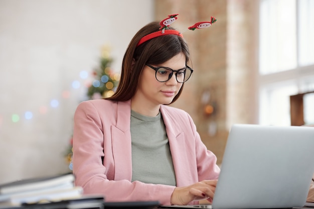 Giovane dirigente d'ufficio piuttosto serio in occhiali, abbigliamento casual intelligente e copricapo natalizio in rete contro abeti decorati