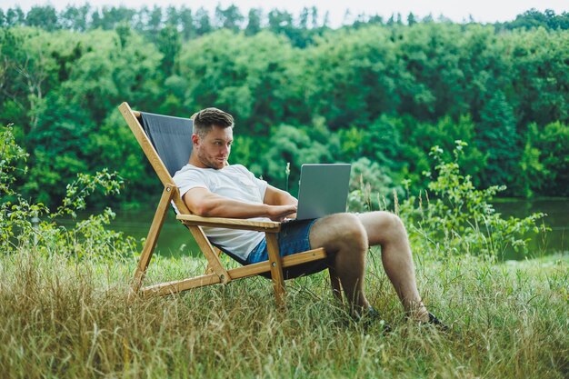 Giovane disteso su una sedia in natura e che lavora con un computer portatile riposando da solo guardando pensieroso Un uomo in viaggio in campagna il concetto di stile di vita delle persone