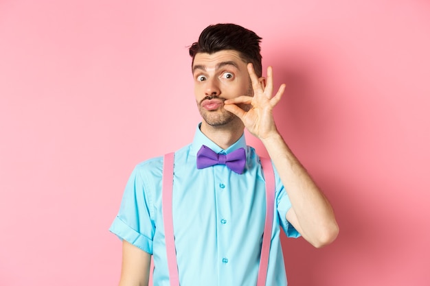 Giovane divertente che tocca i suoi baffi francesi e le labbra arricciate, guardando sciocco alla macchina fotografica, in piedi in cravatta a farfalla e bretelle su sfondo rosa.