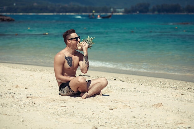 Giovane divertente con occhiali da sole e tatuaggio sul petto sulla spiaggia, le gambe incrociate, con in mano un ananas e fingendo di morderlo; divertimento.