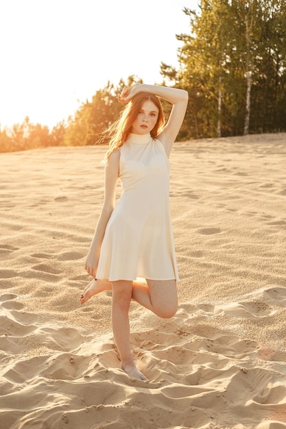 giovane donna a piedi nudi con capelli rossi in abito estivo in piedi sulla sabbia in natura al tramonto