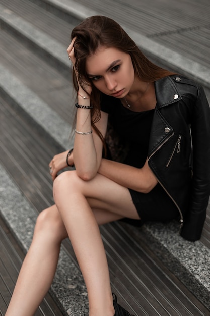 Giovane donna abbastanza alla moda sexy in vestiti neri di autunno alla moda in stivali vintage in pelle sta riposando seduto sui gradini di pietra in strada. Modello urbano bella ragazza moderna si rilassa all'aperto.
