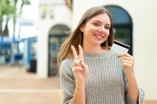 Giovane donna abbastanza bionda che tiene una carta di credito all'aperto che sorride e che mostra il segno di vittoria