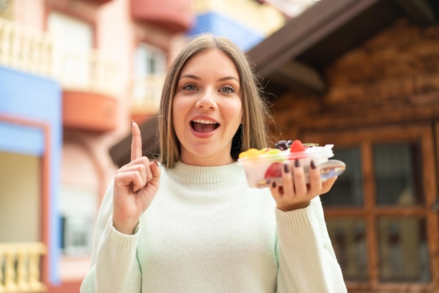 Giovane donna abbastanza bionda che tiene una ciotola di frutta all'aperto che indica una grande idea