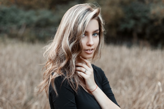 Giovane donna abbastanza carina con trucco naturale con gli occhi azzurri con un sorriso meraviglioso in una maglietta alla moda nera in un campo tra l'erba secca
