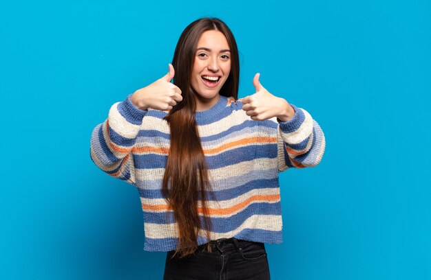 Giovane donna abbastanza casuale che sorride ampiamente felice, positiva, sicura e di successo, con entrambi i pollici in su