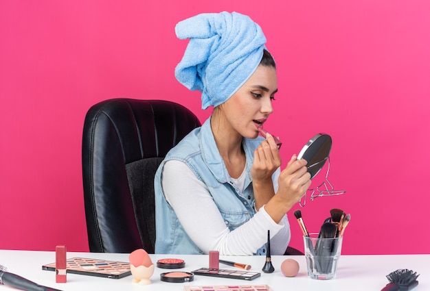 Giovane donna abbastanza caucasica con i capelli avvolti in un asciugamano seduto al tavolo con strumenti per il trucco che tengono e guardano lo specchio applicando il rossetto isolato sulla parete rosa con lo spazio della copia