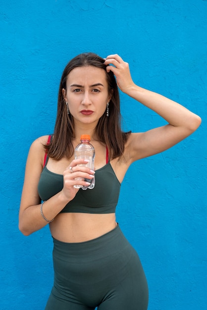 Giovane donna abbastanza esile in abbigliamento sportivo con la bottiglia di acqua della tenuta isolata su fondo blu. Uno stile di vita sano