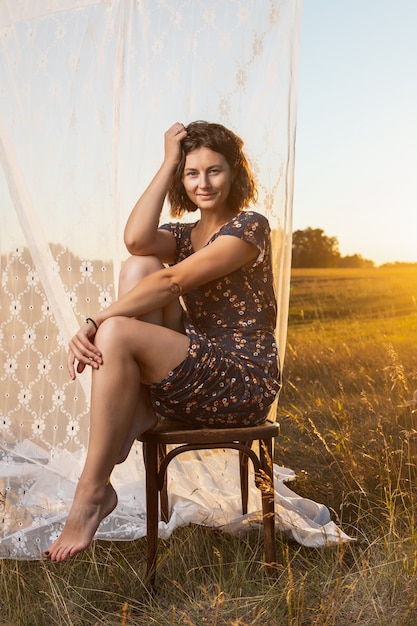 Giovane donna abbastanza fresca che si siede sulla sedia all'aperto nel campo e indossa in abito in background un rack con tende bianche .Concetto di vacanze estive al villaggio e stile dal vivo